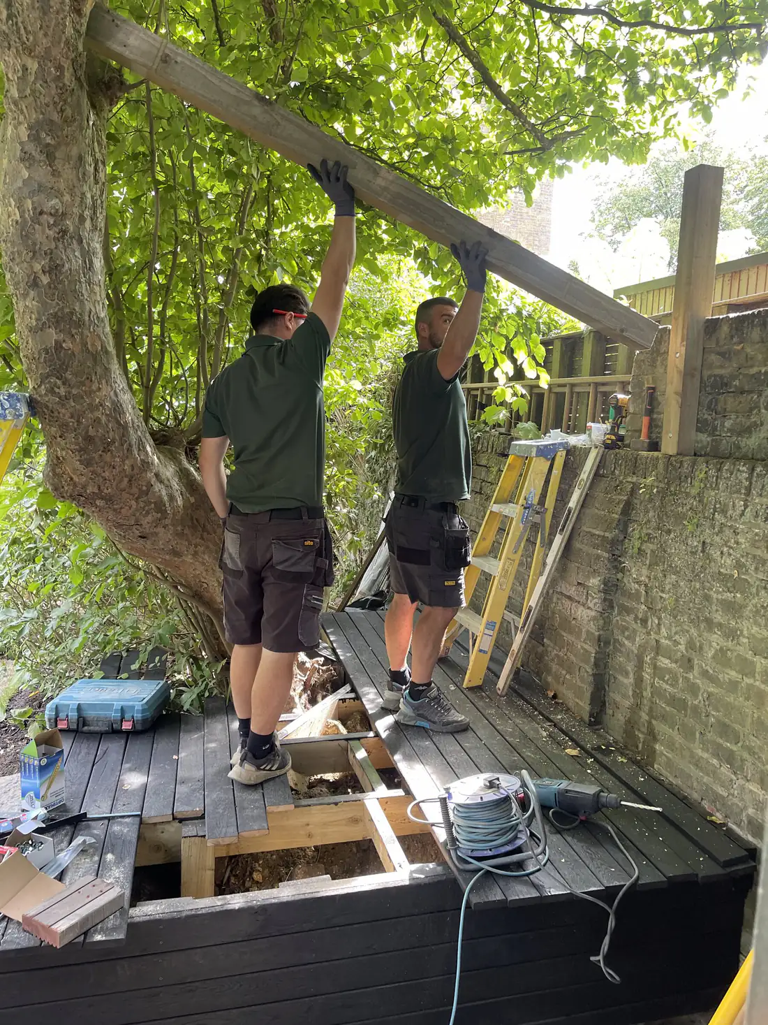 Two people in work polo shirts lifting a timber beam into place in a London garden, drill and stepladder beside them