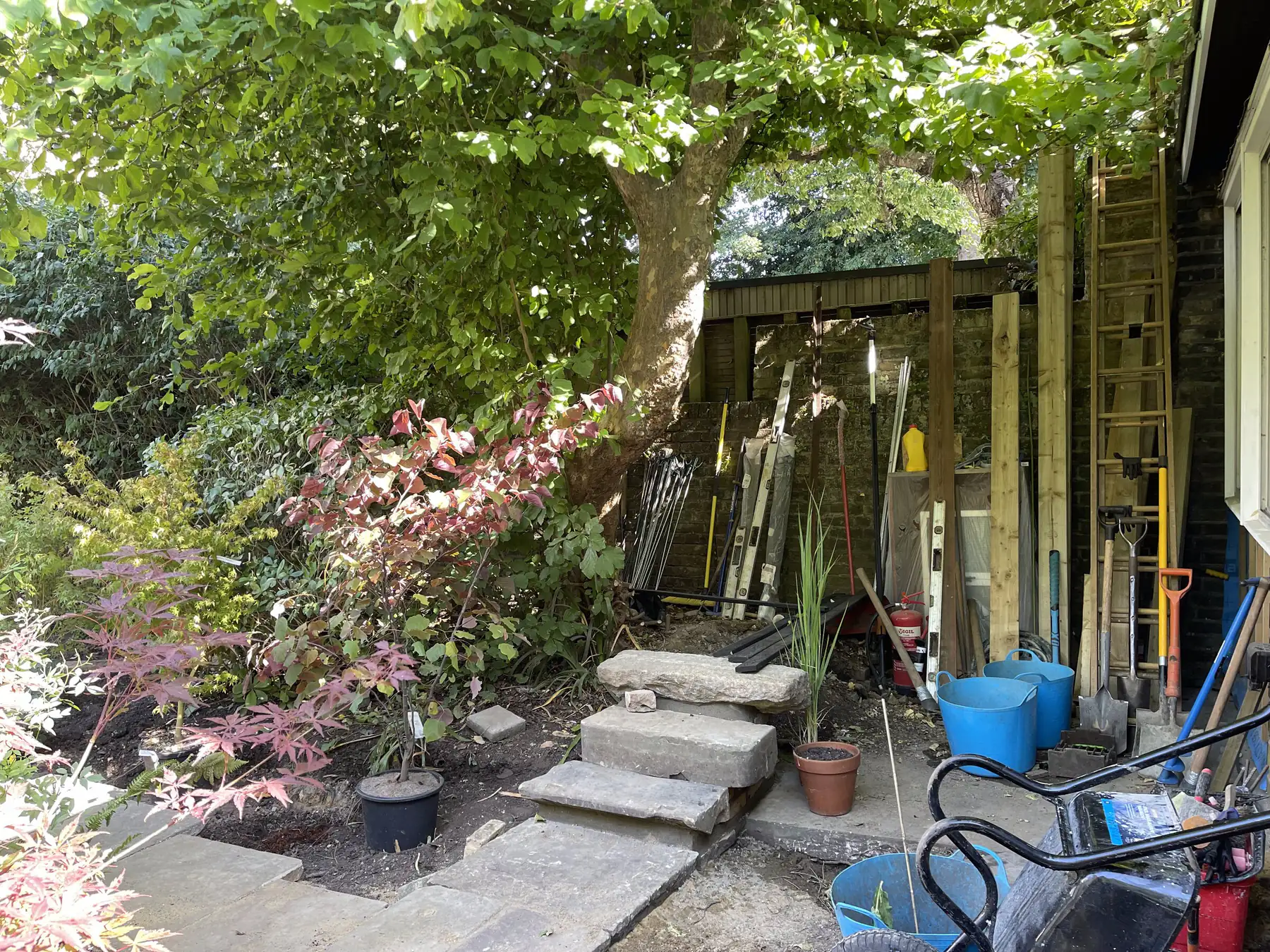 Spades, ladders, trugs and tools stored against a brick wall under a mature tree